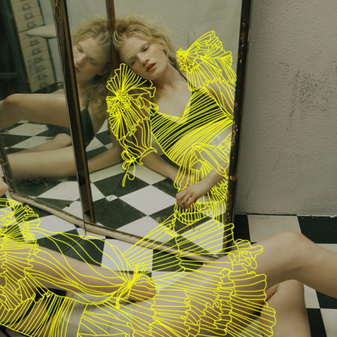 Woman poses by a mirror on a checkered floor, wearing a decorative dress with an illustration overlay in a photo studio setting.