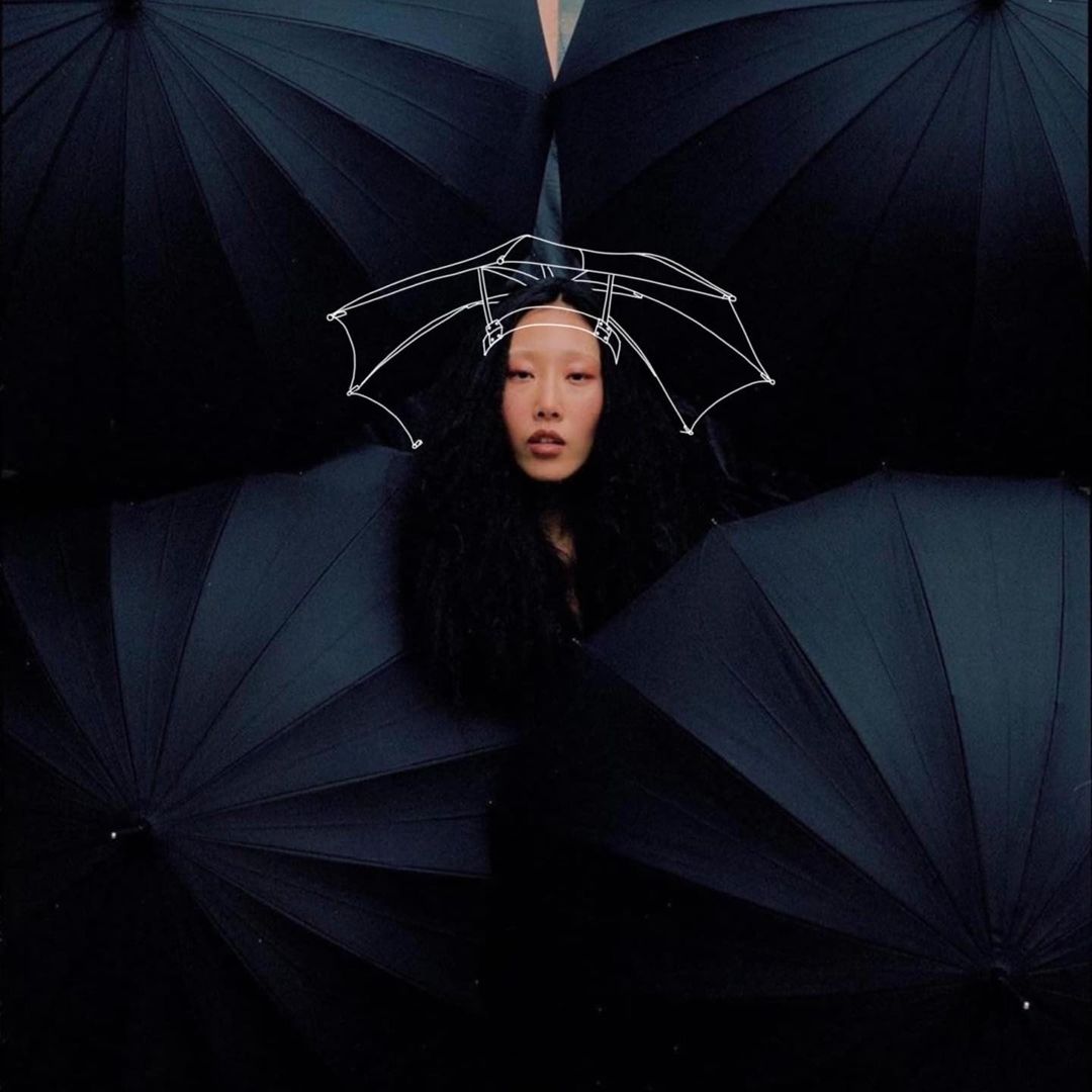 Portrait of an Asian woman framed by several black umbrellas and wearing an umbrella-shaped headpiece. Dark and dramatic lighting.