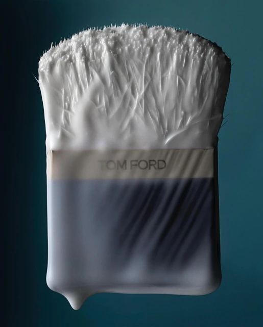 Close-up of a Tom Ford makeup brush encased in clear plastic, bristles at the top, set against a graded teal background.