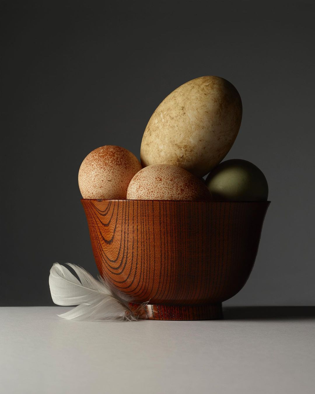 Still life shot of a wooden bowl filled with several eggs of different colors and sizes, and a white feather on a white surface.