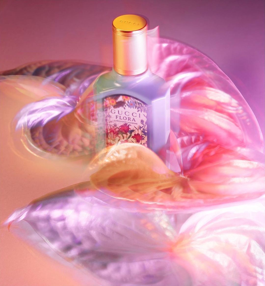 Gucci Flora perfume bottle with gold lid on a pink surface with a blurred floral background, creating a luxurious and soft aesthetic.