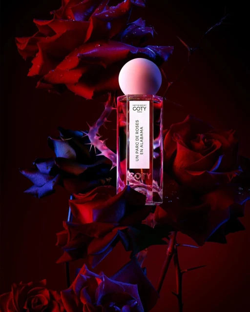 Elegant product shot of a Coty perfume bottle with a round white cap, surrounded by vibrant red and purple roses, with a dark background.