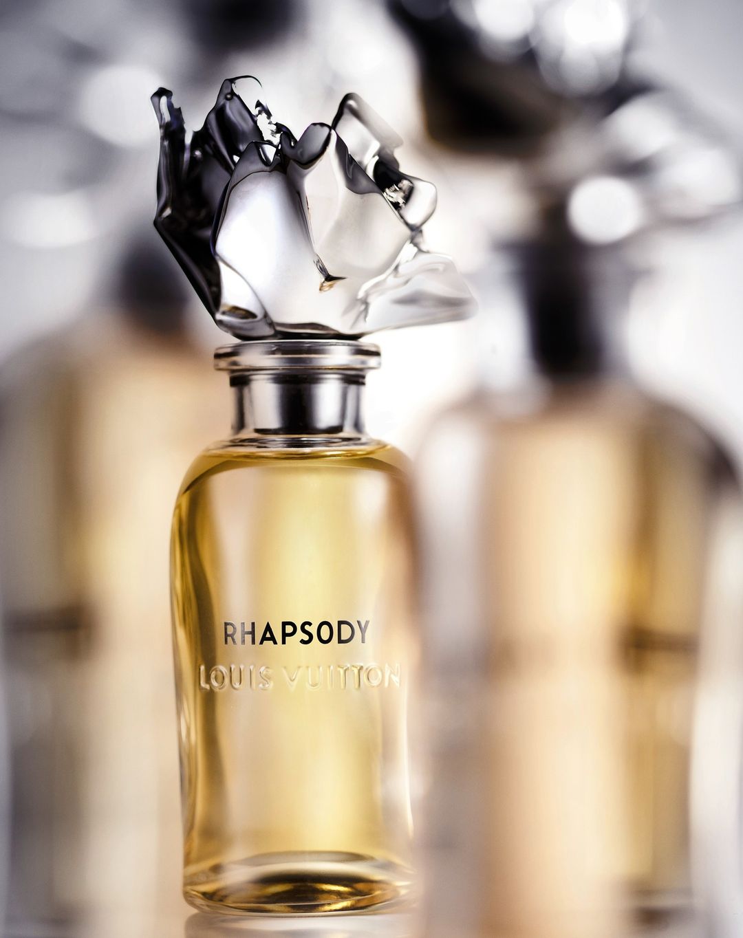 Close-up studio shot of a Louis Vuitton Rhapsody perfume bottle with a clear glass flacon and silver top, against a blurred background.