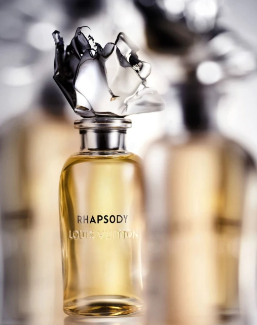 Close-up studio shot of a Louis Vuitton Rhapsody perfume bottle with a clear glass flacon and silver top, against a blurred background.
