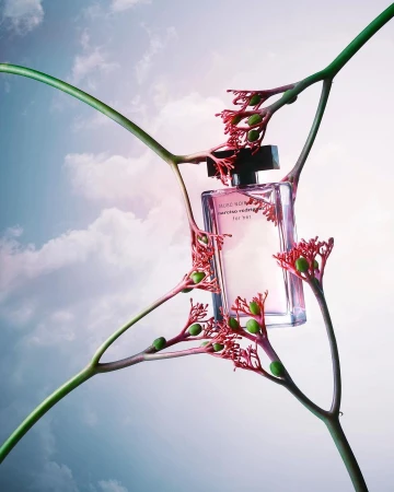 Glass perfume bottle centered in a composition with green stems and red buds against a background of blue sky and soft white clouds.