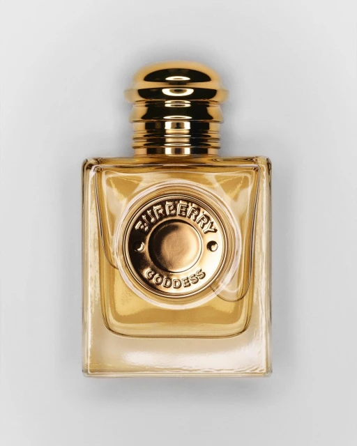Gold-toned Burberry Goddess perfume bottle in a square shape with a circular emblem against a neutral background in a studio setting.