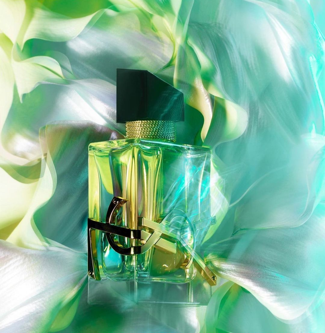 Studio shot of a green glass perfume bottle with a black lid against a blurred backdrop of swirling light green and white fabric.