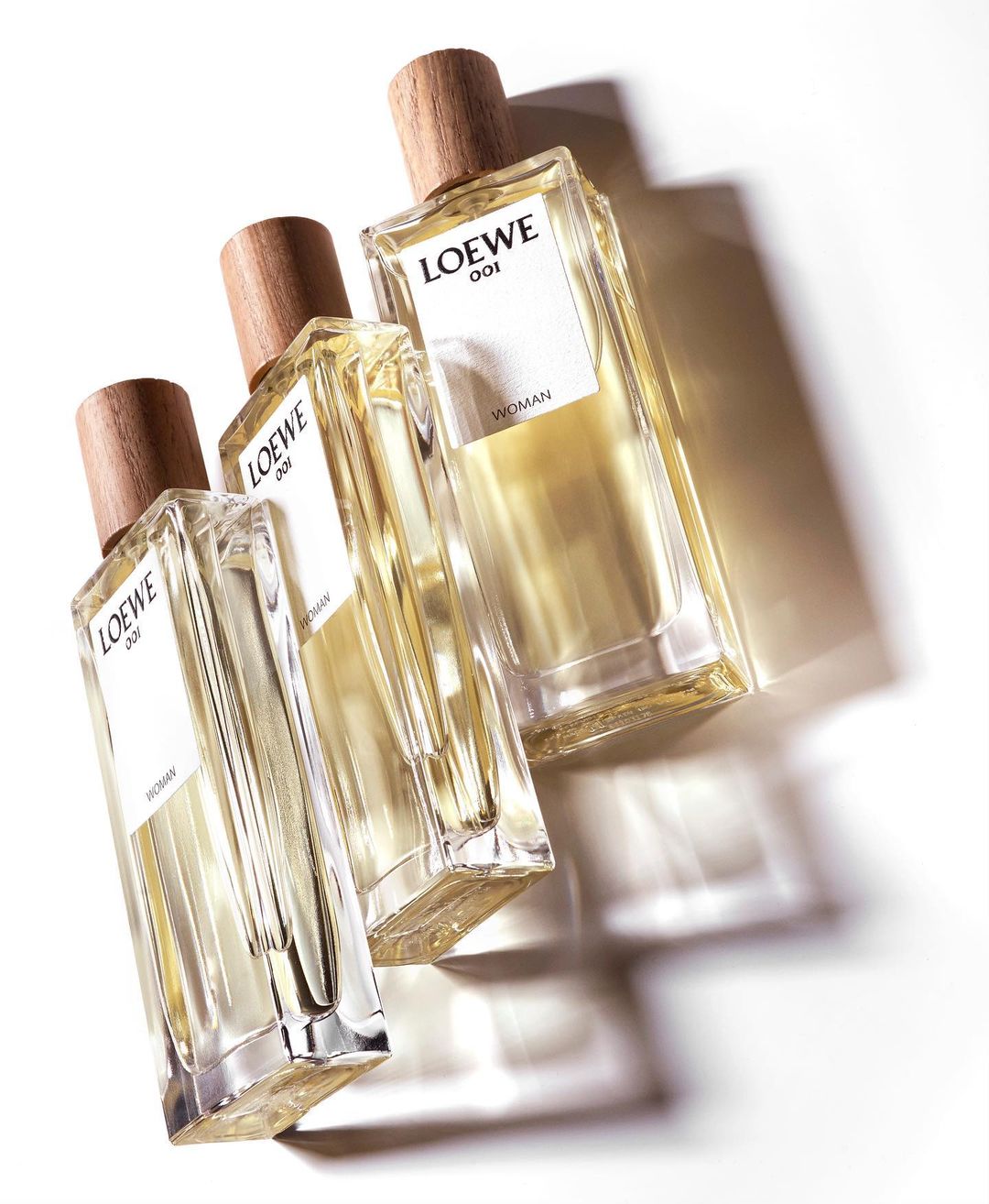 Three Loewe 001 Woman perfume bottles are angled on a white surface casting shadows, product shot, beauty and fashion concept.