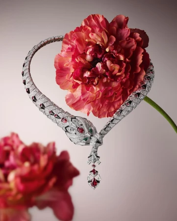 Close-up of a silver snake necklace featuring diamonds, rubies, and emeralds, intertwined with a pink ranunculus on a pale gradient background.