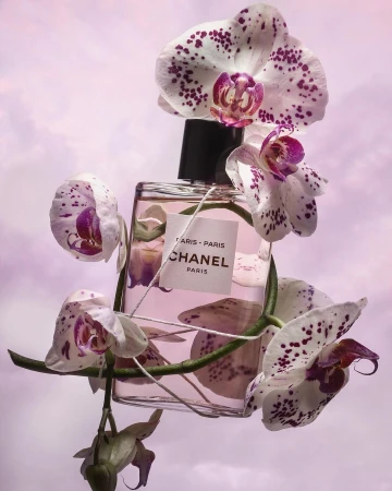 Still life shot of a Chanel perfume bottle encircled by white and purple orchid flowers against a dreamy soft purple backdrop.