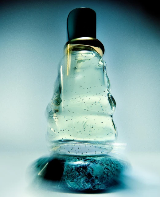 Clear glass perfume bottle with a black cap rests on a teal stone base, set against a soft, gradient blue background.