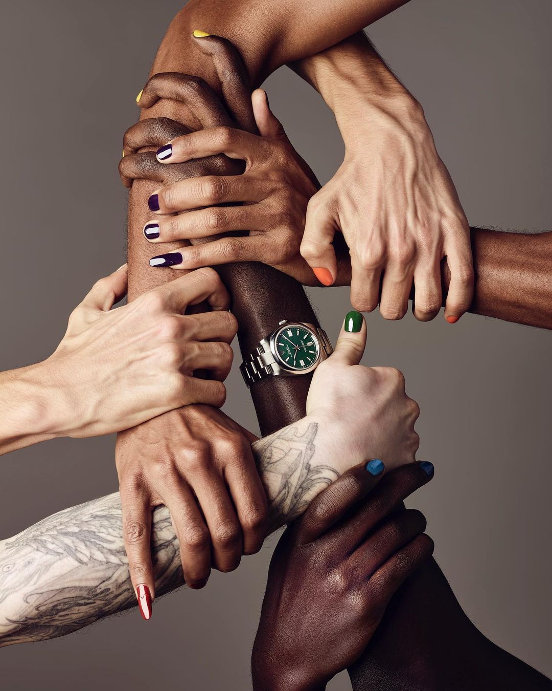 Diverse hands join together, one wearing a green-faced watch, displaying unity and inclusivity in a neutral-toned setting, studio shot.