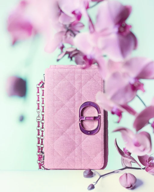 Close-up of a pink quilted handbag with chain strap, featuring a metal logo detail, arranged with pink orchids against a soft teal background.