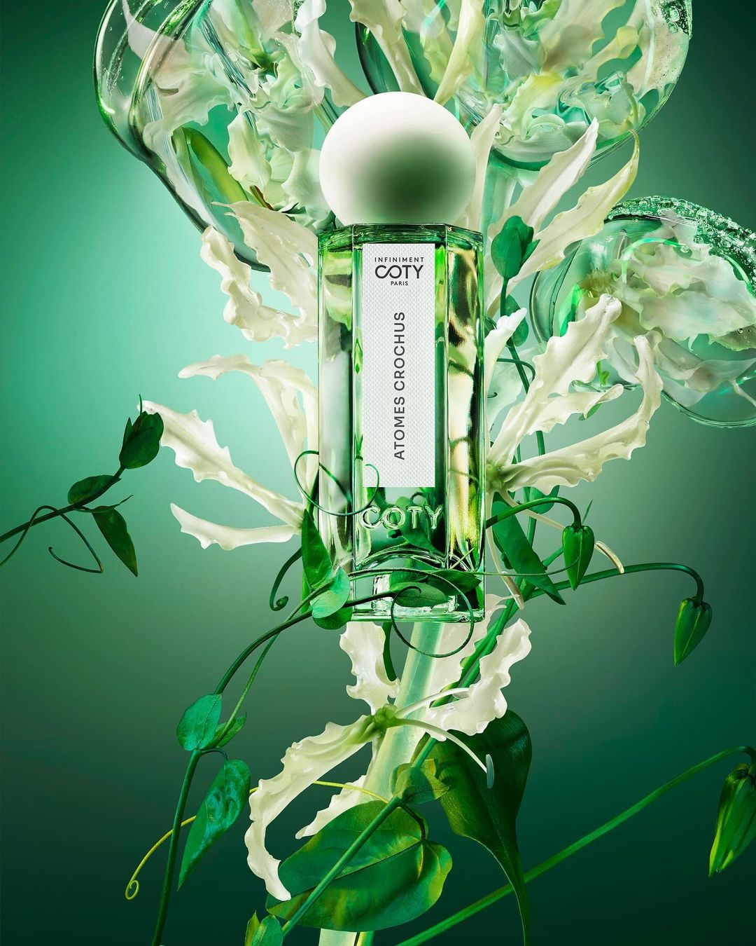 Coty Atomes Crochus perfume bottle with a white round cap surrounded by white Gloriosa superba lilies and green leaves against a green background.