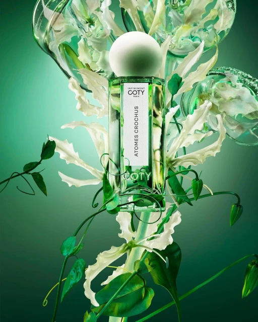 Coty Atomes Crochus perfume bottle with a white round cap surrounded by white Gloriosa superba lilies and green leaves against a green background.
