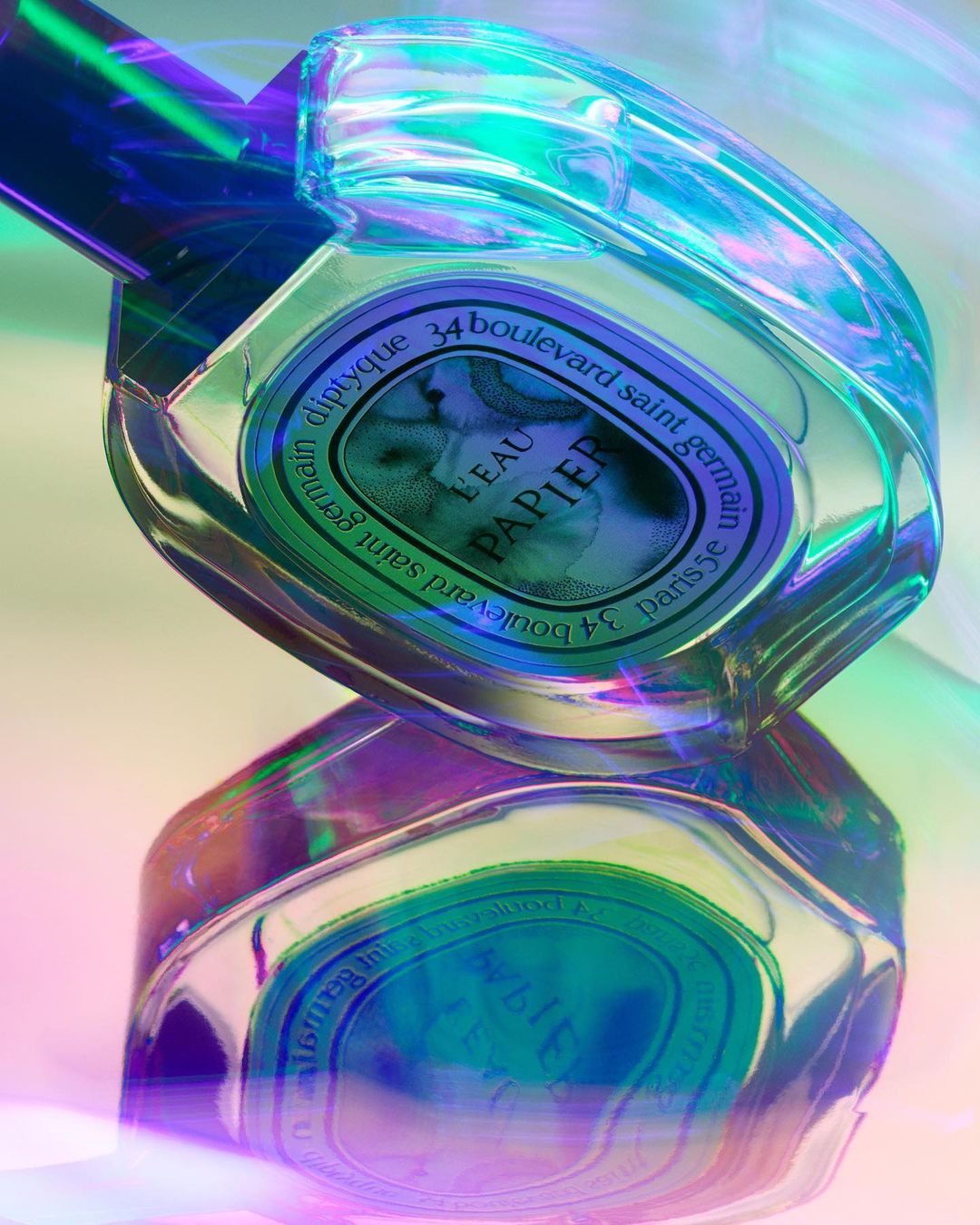 Close-up studio shot of a Diptyque Eau Papier perfume bottle reflecting on a glossy surface with a holographic gradient light effect.
