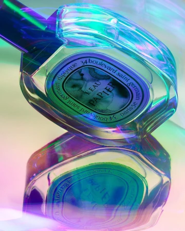 Close-up studio shot of a Diptyque Eau Papier perfume bottle reflecting on a glossy surface with a holographic gradient light effect.