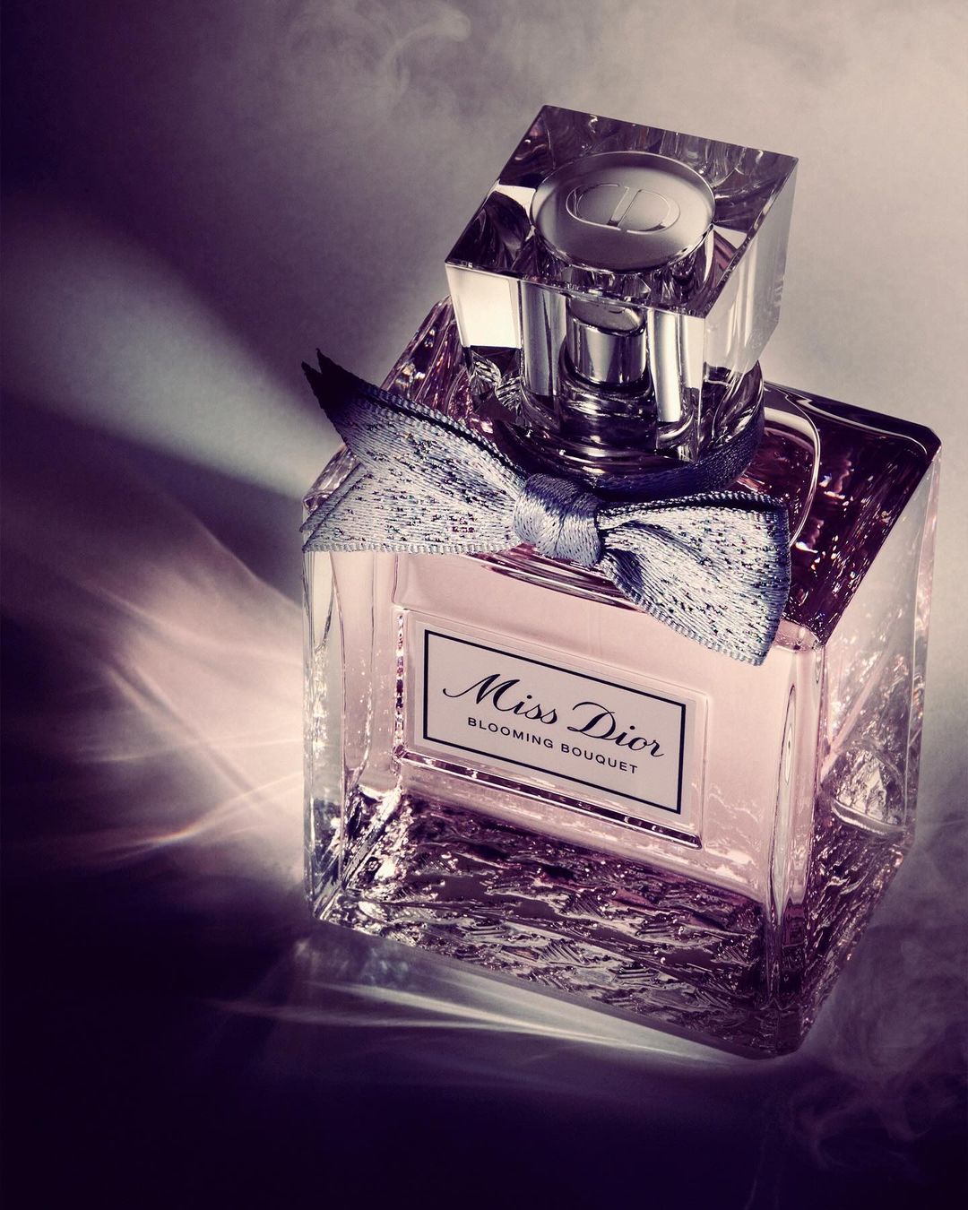 Crystal bottle of Miss Dior Blooming Bouquet perfume with a silver bow sits on a patterned surface with smoke and dramatic lighting.