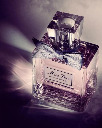 Crystal bottle of Miss Dior Blooming Bouquet perfume with a silver bow sits on a patterned surface with smoke and dramatic lighting.