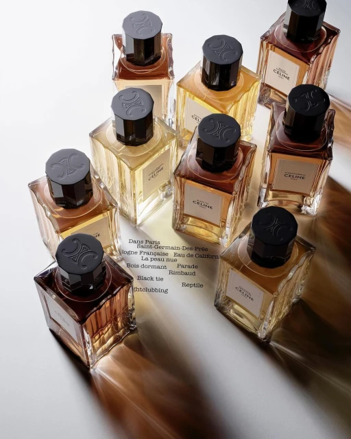 Multiple square glass perfume bottles from Celine, filled with golden and brown liquids, arranged on a white surface with shadows.