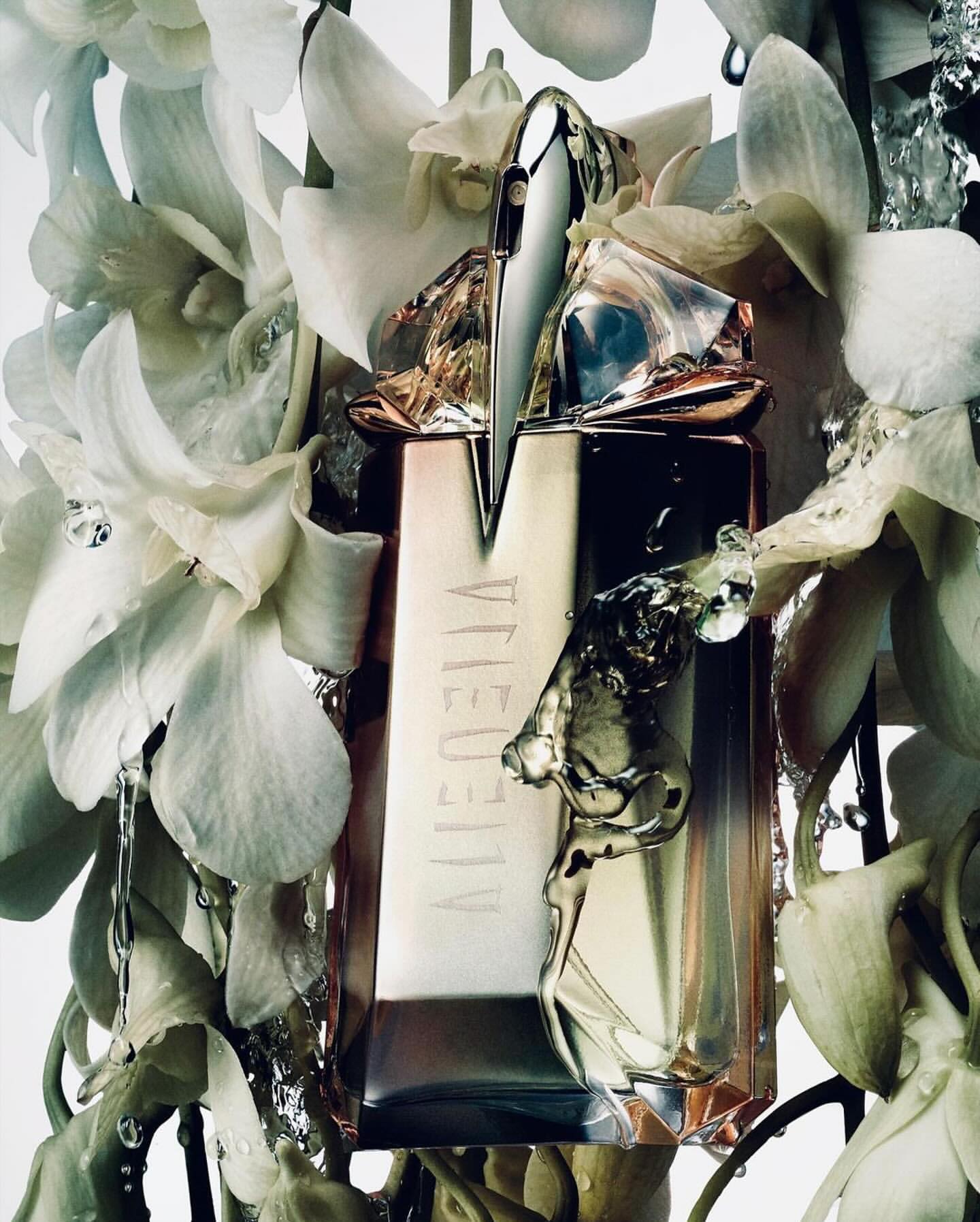 Close-up studio shot of a sculptural perfume bottle surrounded by delicate white orchid blossoms and water droplets on a white background.