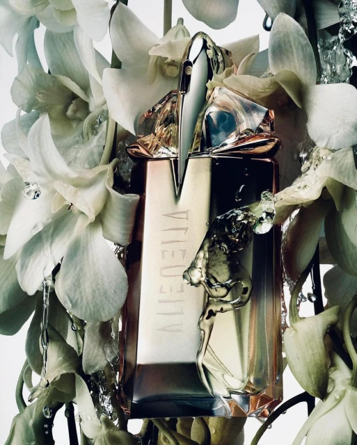 Close-up studio shot of a sculptural perfume bottle surrounded by delicate white orchid blossoms and water droplets on a white background.