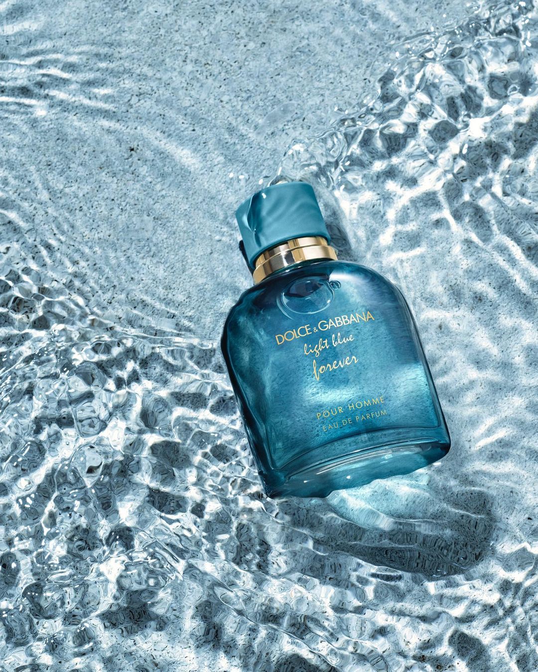 A close-up product shot showcases a light blue Dolce & Gabbana men's cologne bottle submerged in clear, rippling water against a neutral background.