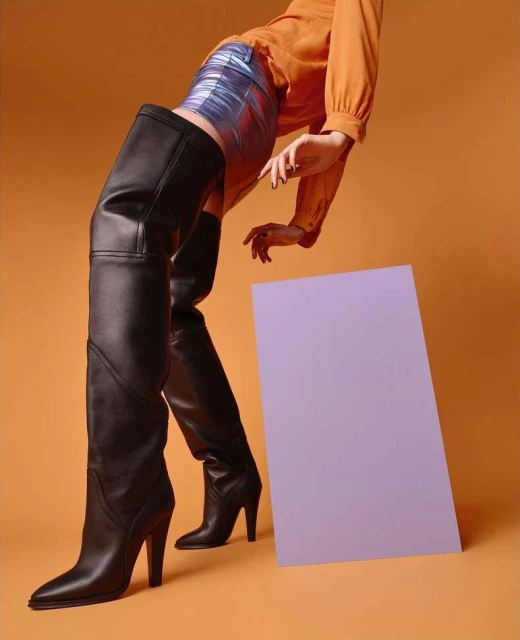 Fashion editorial shot of a woman wearing thigh-high black boots, a short blue metallic skirt, and an orange blouse against an orange background.
