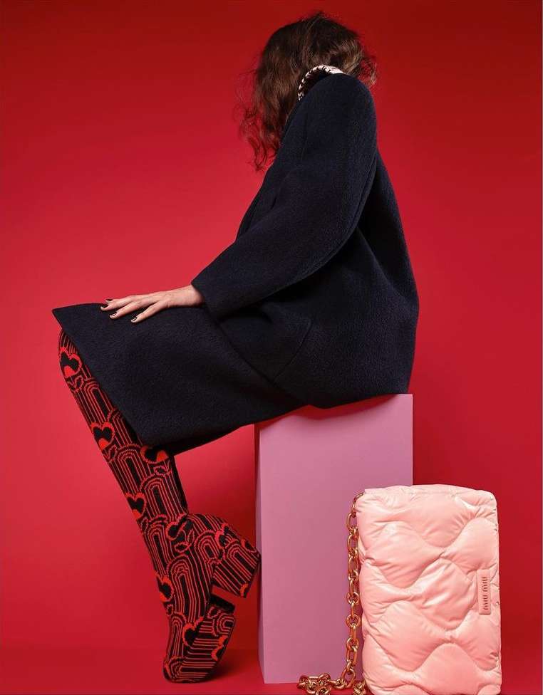 A fashion shot featuring a woman in a dark coat and patterned tights, seated on a pink cube with a light pink purse nearby against a red backdrop.