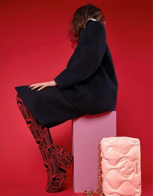 A fashion shot featuring a woman in a dark coat and patterned tights, seated on a pink cube with a light pink purse nearby against a red backdrop.