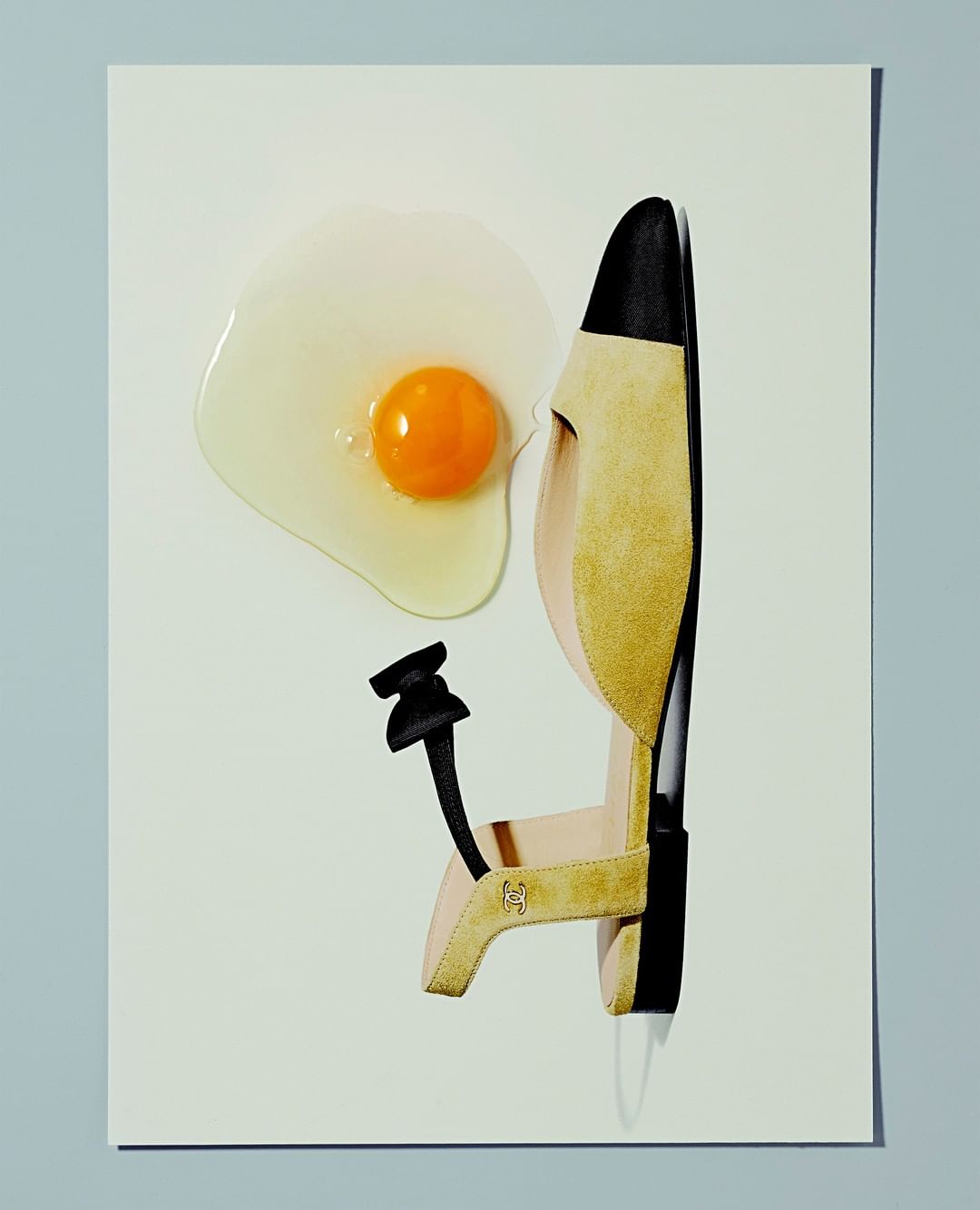 Fashion still life: beige and black sandal arranged next to a fried egg on a white surface with a light blue background.
