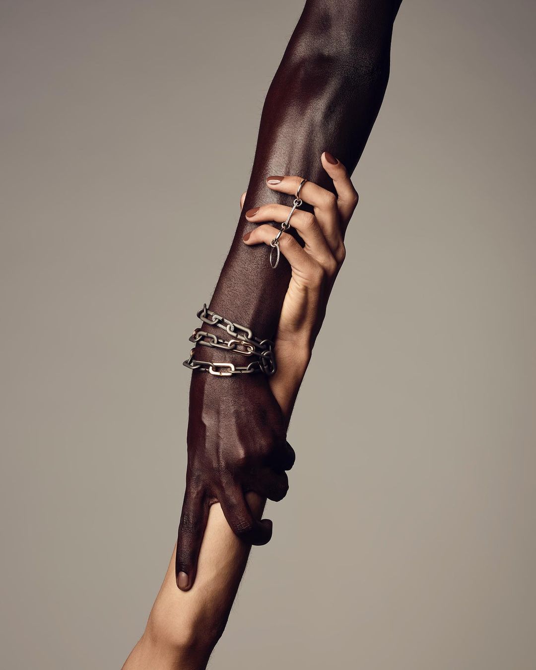Close-up studio shot of two adult human arms with different skin tones, one with rings, one with a chain bracelet, holding hands.