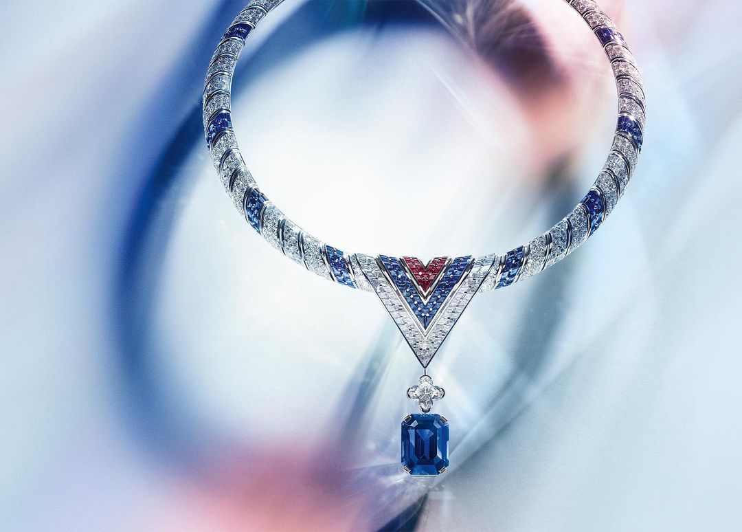 Elegant diamond and sapphire necklace featuring a V-shaped pendant with a large blue gemstone, set against a blurred, soft-colored background.