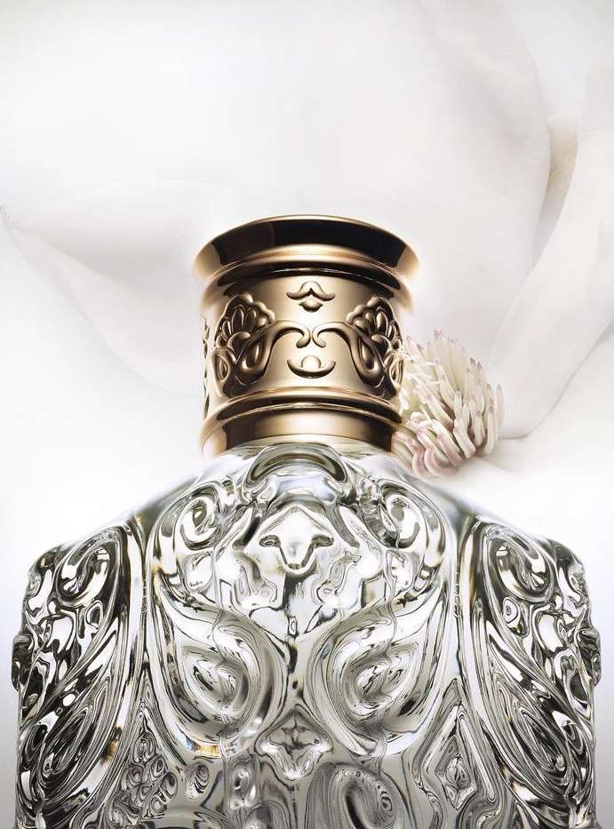 Close-up of a decorative clear glass perfume bottle with gold ornate cap, and soft white flower in the background on a white backdrop.
