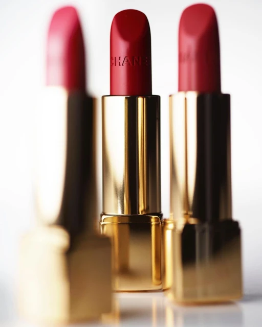 Close-up of three gold Chanel lipstick tubes with red lipstick against a gradient white background; beauty and fashion product photography.