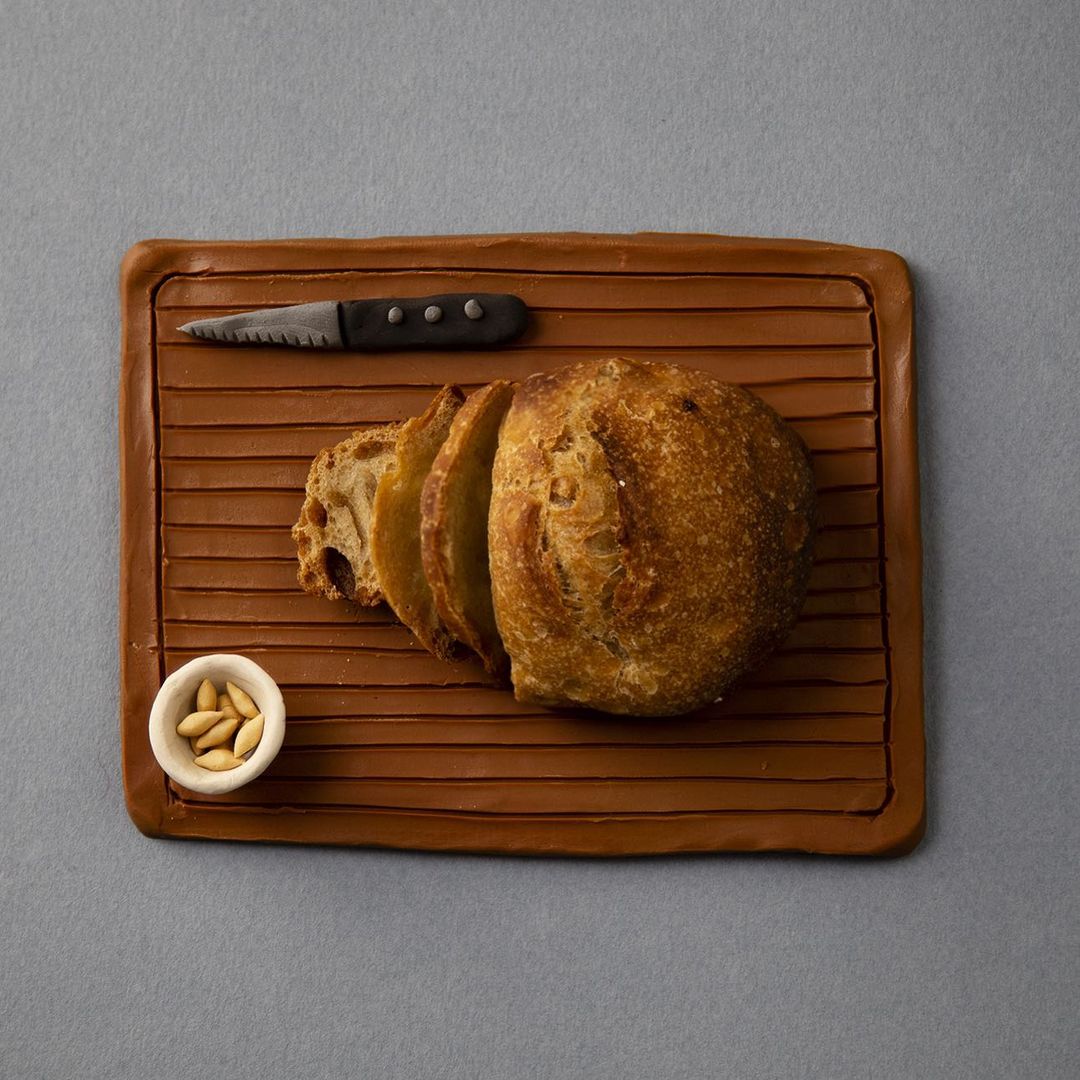 Overhead shot of sliced sourdough bread and knife resting on a wooden serving board with a ramekin of pine nuts, tabletop setting.