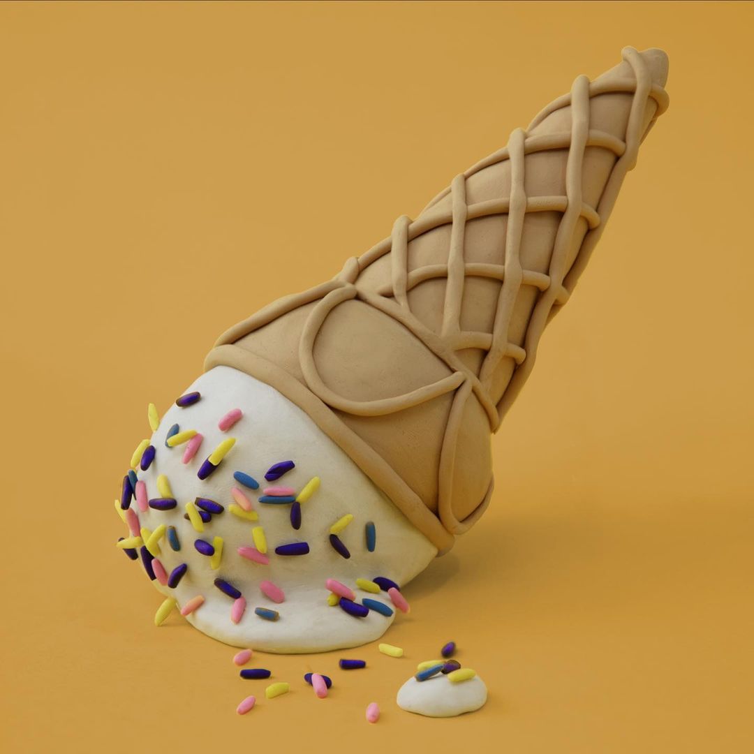 Stylized studio shot of an ice cream cone on its side, sprinkles scattered around the fallen scoop against a solid yellow background.