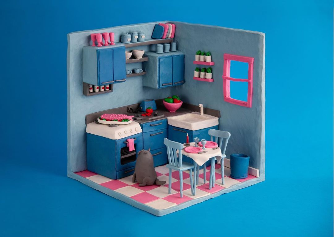 Quirky stop-motion kitchen diorama: Blue cabinets, pink checkered floor, toy cat, and dinner table against a solid blue backdrop.