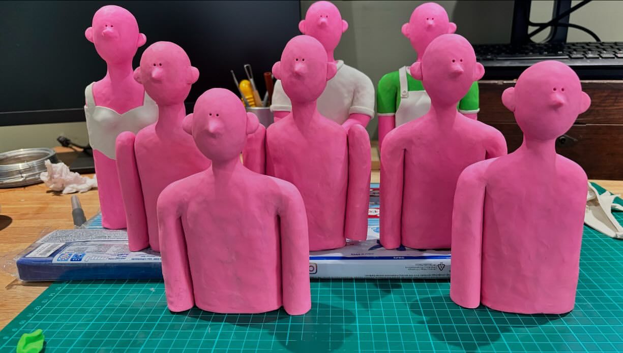 Nine pink clay figures are arranged on a work surface with a green cutting mat, some wearing clothing, in an indoor studio setting.