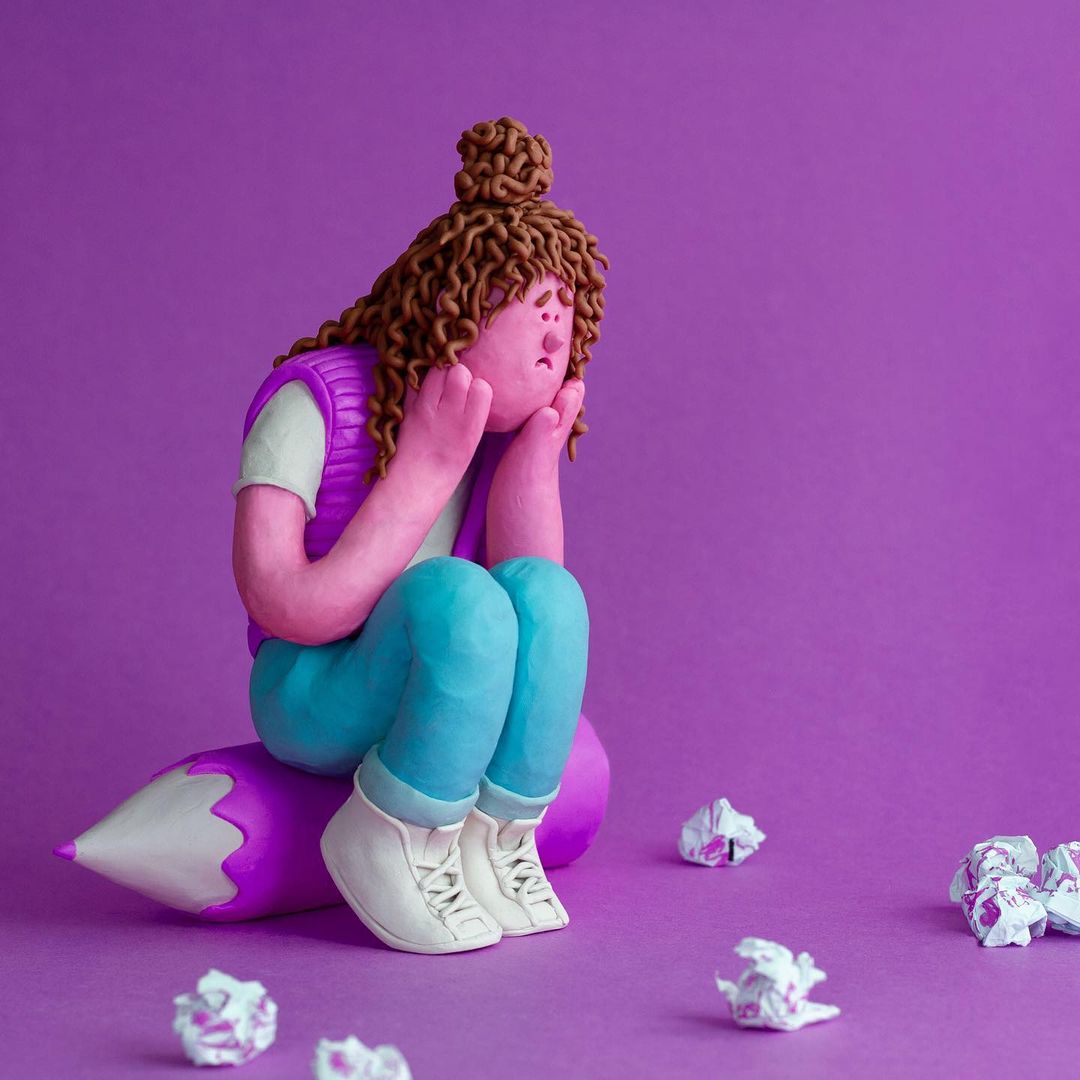 Claymation figurine of a woman sitting atop a purple pencil surrounded by crumpled papers, set against a purple background.