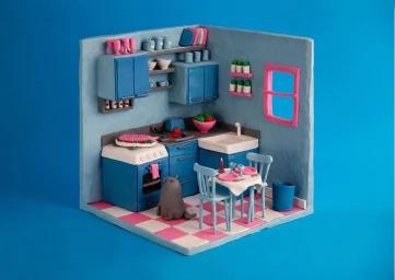 Handcrafted miniature clay kitchen diorama with blue cabinets, checkered pink floor, and a small cat sitting on the floor by Inès Pagniez