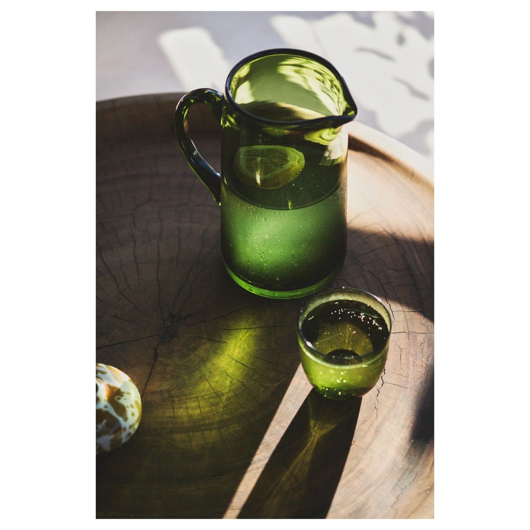 Green glass pitcher with water, lemon slice, and a glass on a round wooden tray in natural light creates a refreshing, vibrant composition.