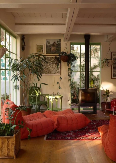 Living room interior with orange modular sofas, a fireplace, indoor plants, and natural light streaming through the large windows.