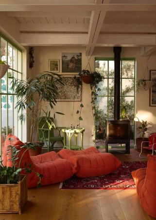 Living room interior with orange modular sofas, a fireplace, indoor plants, and natural light streaming through the large windows.