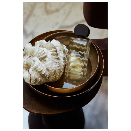 Product shot of a clear glass perfume bottle with black cap, next to a textured white shell, sitting in a gold bowl on a brown pedestal.