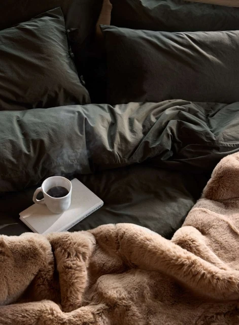 Cozy bedroom scene with a steaming mug on a book atop an olive green duvet, accented by a textured faux fur throw blanket.