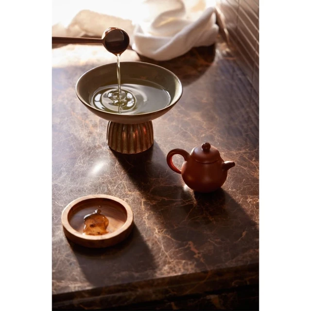 Close-up of liquid being poured from a wooden spoon into a green bowl, along with a small wooden teapot on a marble surface.