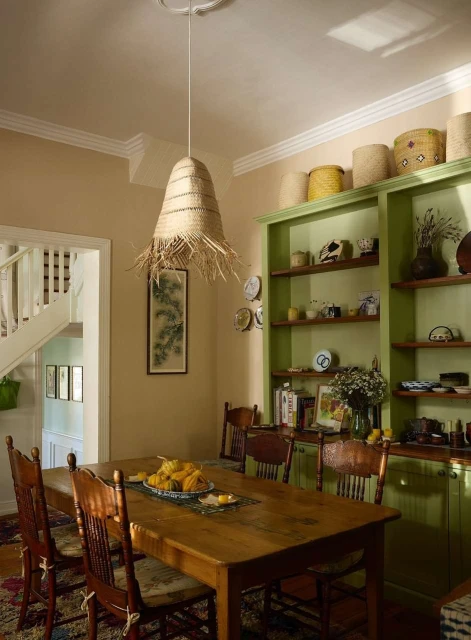 Dining room interior with wooden table, chairs, straw pendant light, and green shelving unit filled with decorative items and dishware.