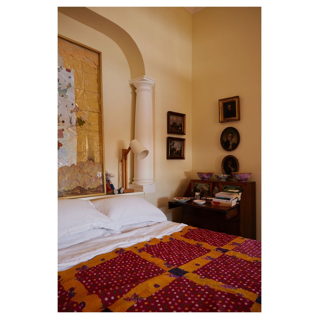 Bedroom interior with a bed covered in a patterned quilt, a desk with books, and artwork on the walls, creating a cozy and artistic space.