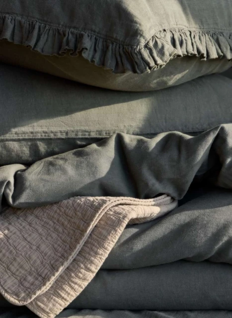 Close-up of green linen bedding, including a pillow with ruffles, and a folded beige throw blanket creating a cozy, inviting texture.